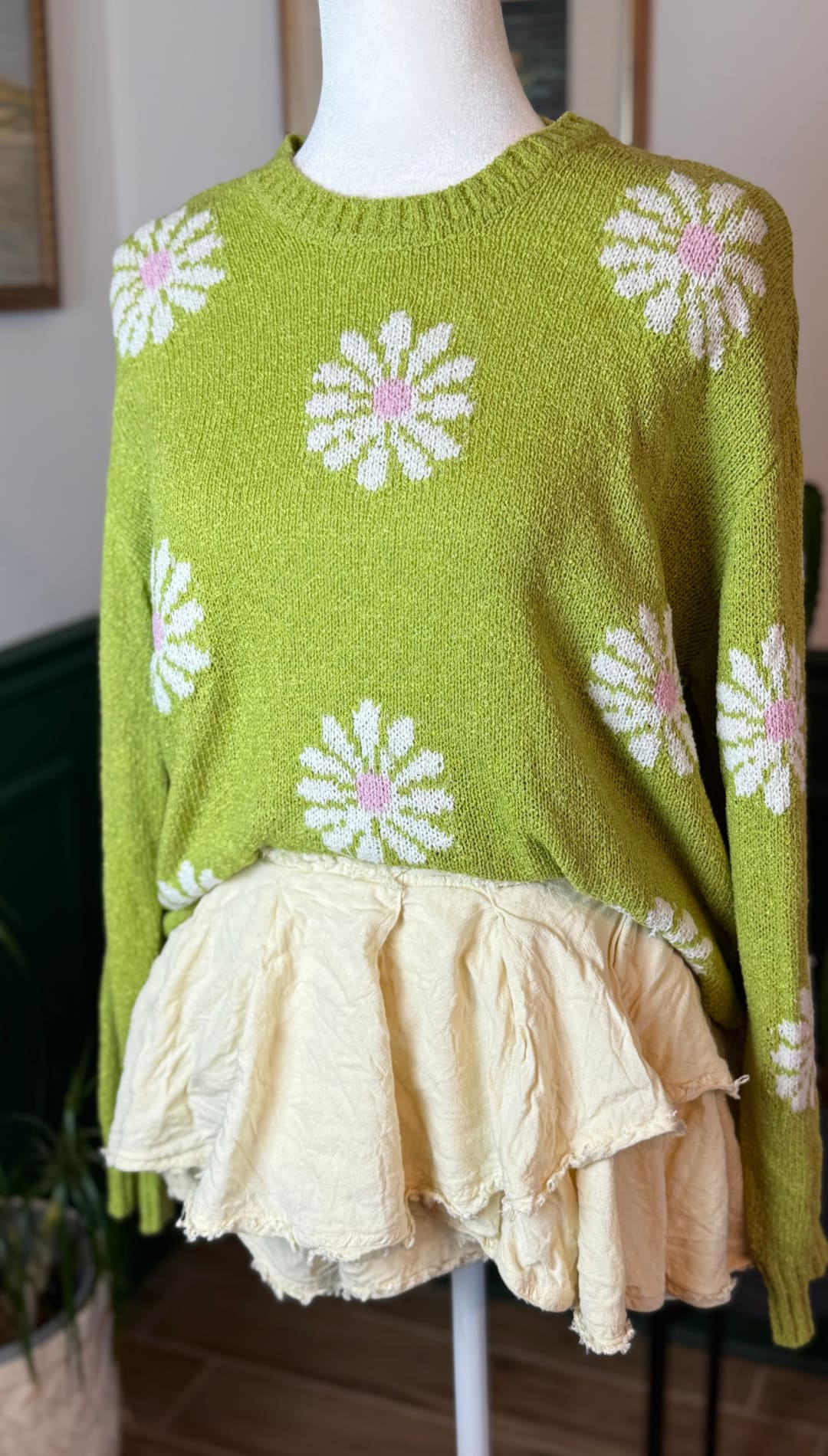 A lime green knit sweater with white and pink daisy patterns, featuring a ruffled cream-colored skirt overlay.