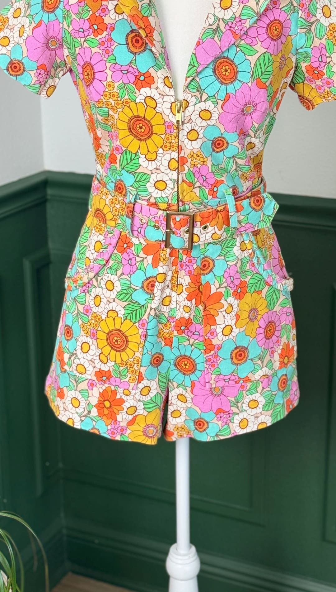 Vibrant floral print romper with short sleeves, a belted waist featuring a gold-toned buckle, and pockets on the sides.