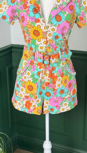 Vibrant floral print romper with short sleeves, a belted waist featuring a gold-toned buckle, and pockets on the sides.
