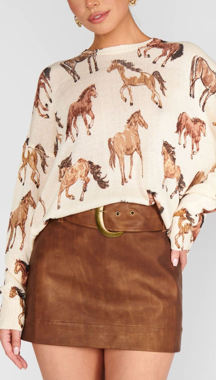 A cream-colored long-sleeve sweater featuring a repeating brown horse print, paired with a brown leather mini skirt cinched by a wide belt with a gold buckle.