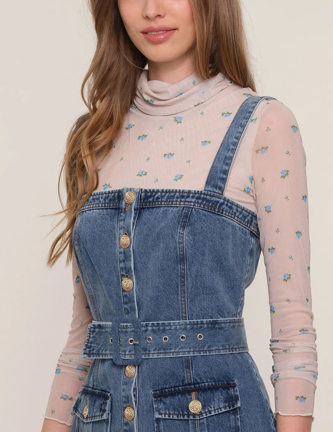 A woman wearing a denim overall dress over a sheer floral long-sleeve top.