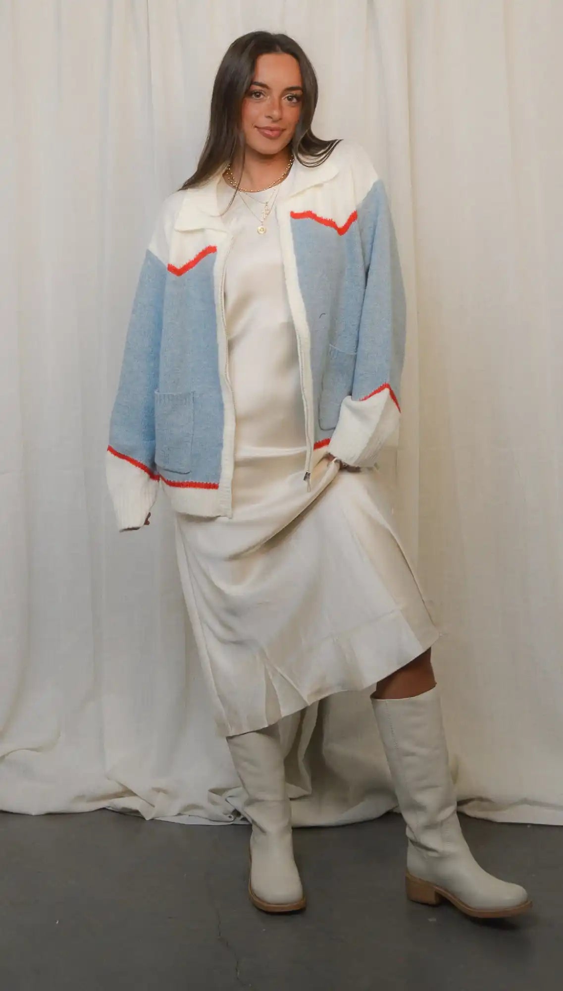 A woman modeling a light blue and white color block cardigan with red trim, a cream-colored midi dress, and tall white boots.
