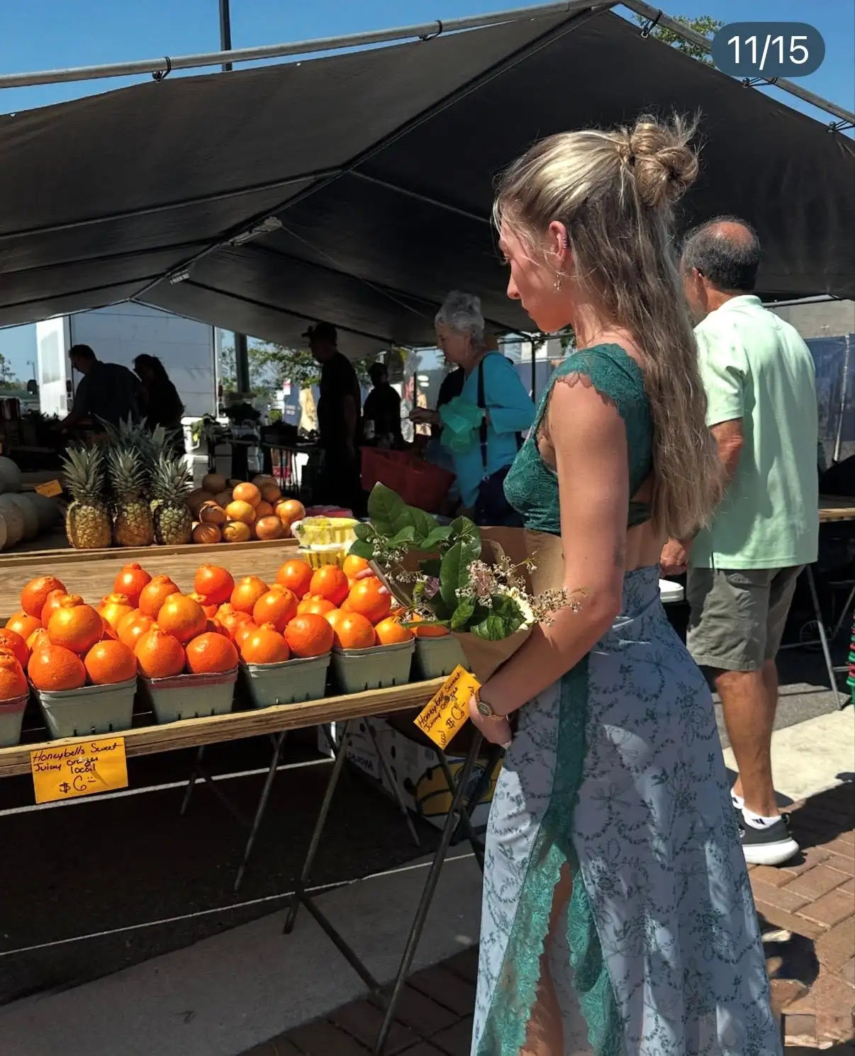 A woman in a teal lace crop top and light blue patterned skirt stands at an outdoor market stall, holding a bouquet of flowers.