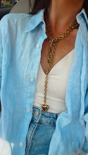 Gold-toned chain necklace with a heart-shaped pendant, layered over a light blue button-up shirt.