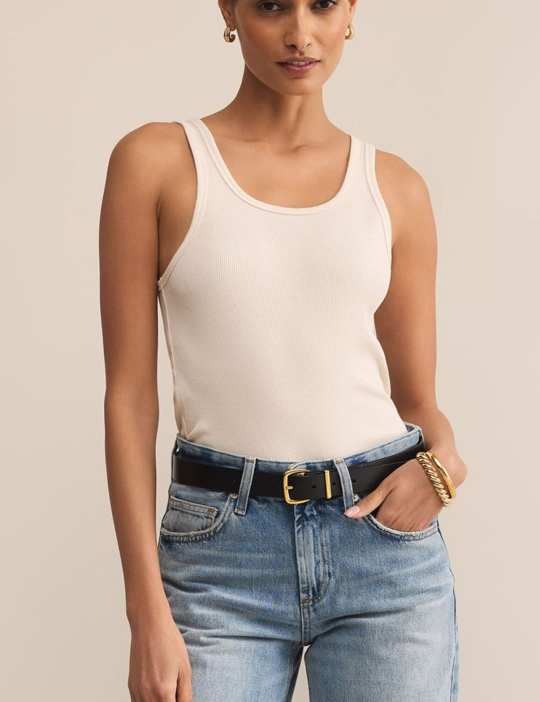 A woman in a cream ribbed tank top, light wash jeans, and a black belt.