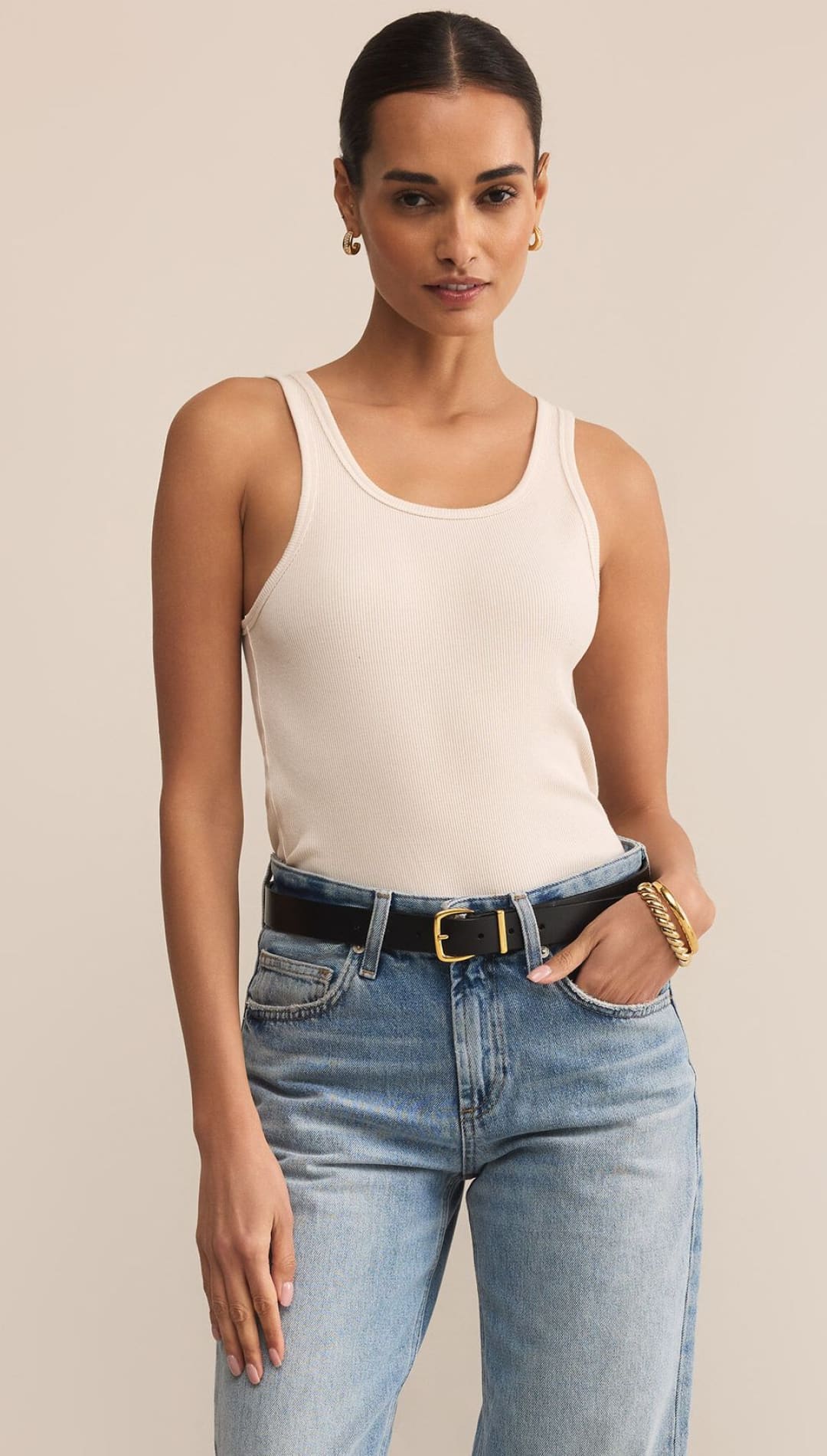 A woman in a cream ribbed tank top, light wash jeans, and a black belt.