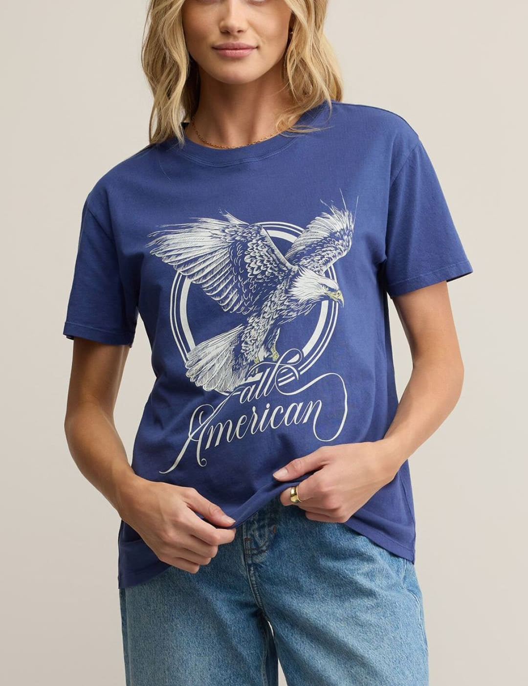 A woman wearing a blue t-shirt with a white graphic of an eagle and the words ’all American’ and light wash blue jeans.