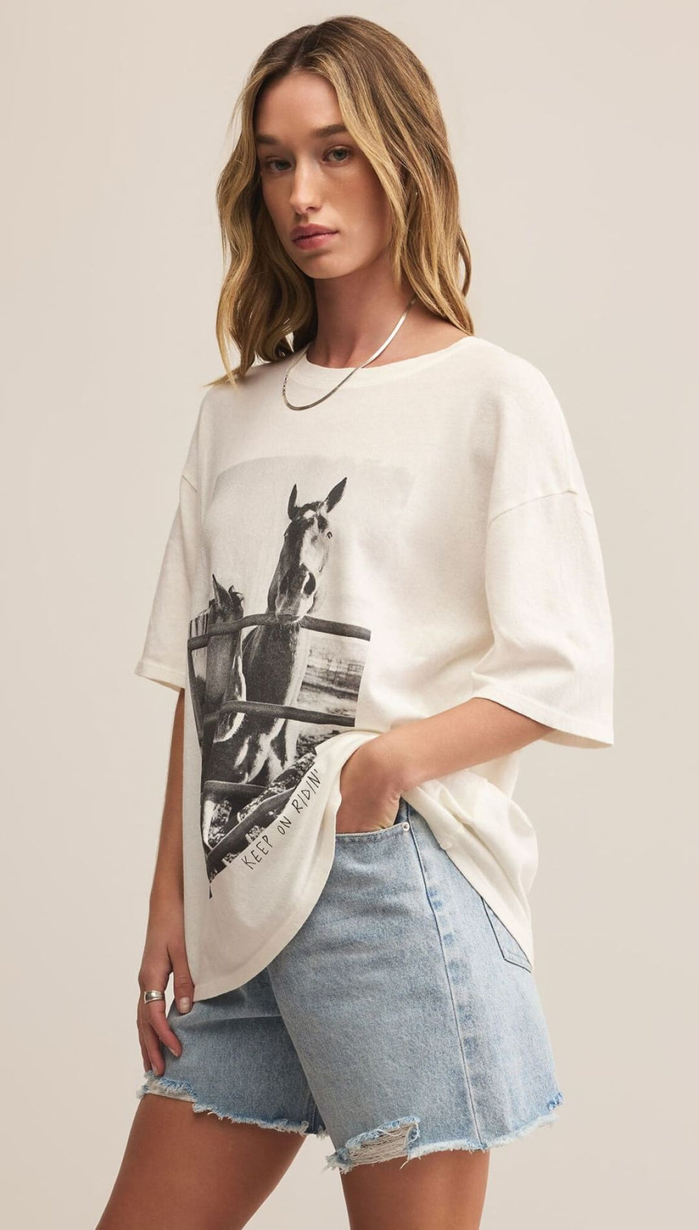 A woman wearing a cream-colored t-shirt with a black and white horse graphic and light-wash denim shorts.