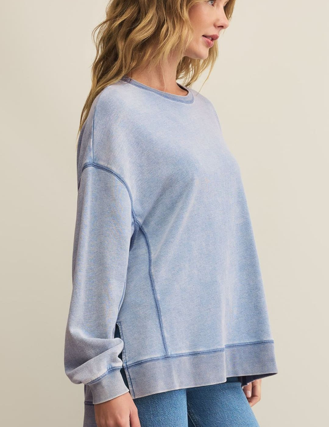 A woman wearing a light blue, long-sleeved crewneck sweatshirt with visible seams and light-wash blue jeans.