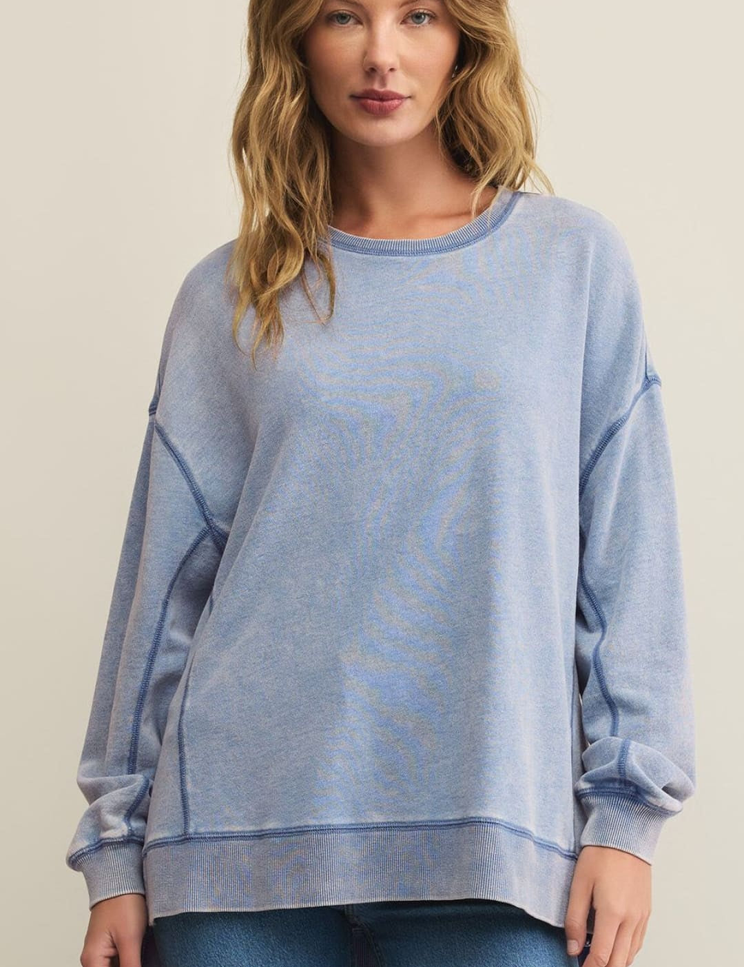 A woman modeling a relaxed-fit, acid-wash blue crewneck sweatshirt with visible stitching and blue jeans.