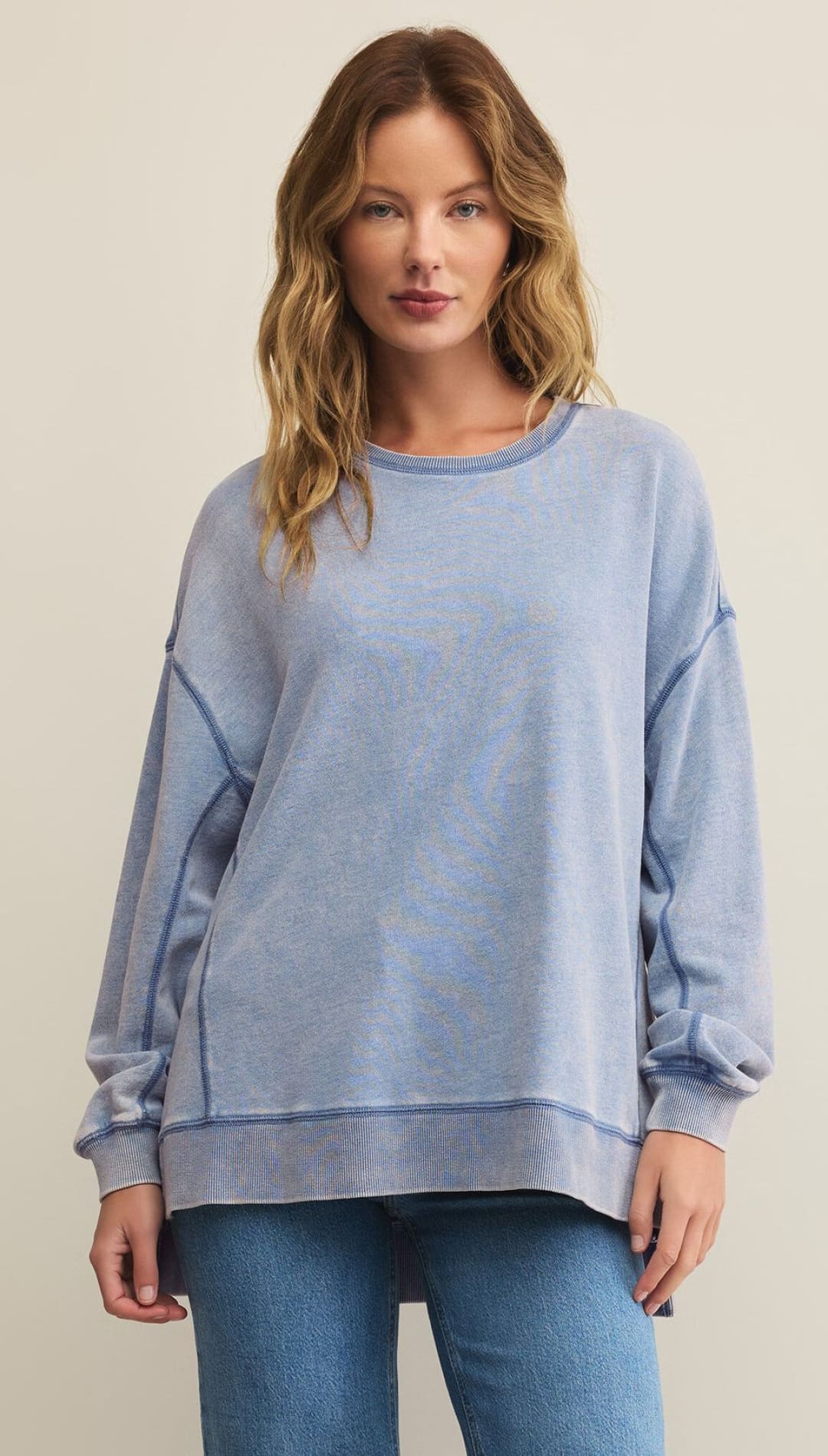 A woman modeling a relaxed-fit, acid-wash blue crewneck sweatshirt with visible stitching and blue jeans.